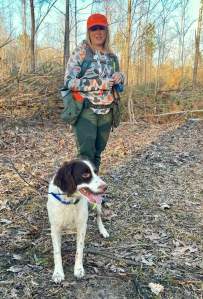 A person wearing an orange hat and camouflage clothing stands on a forested path beside a white dog with brown markings.